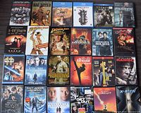 Top view of 25 DVD and Blu-Ray movies laid out in a grid showing front cover artwork including titles such as The Expendables, Bad Boys II, Inkheart, Ocean's Thirteen, Thor, Faster, Hero, Indiana Jones, Sherlock Holmes, Star Wars, Star Trek, The Karate Kid, Rambo, and others.