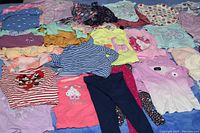 Multiple toddler girls tops spread out showing various colors and patterns, including cartoon prints and stripes.
