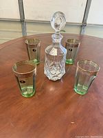 Full set showing four green-tinted crystal glasses with gold Greek key pattern trim around the rims and a clear crystal decanter with stopper on a wooden table.