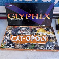 Closed boxes of Glyphax and Cat-Opoly games stacked together showing front cover art and titles.