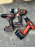 Three used Black & Decker cordless drills shown from above on concrete surface with batteries attached.