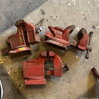 Three red vintage bench vises arranged on concrete floor, showing front and side angles with legible markings.