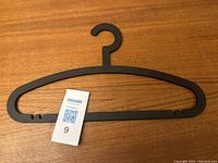 Black plastic IKEA hanger laid on wooden surface, showing full front view with MaxSold tag number 9