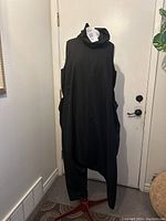 Full view of black sleeveless one-piece jumpsuit on a mannequin against a door background, showing cowl neck and harem pant style legs.