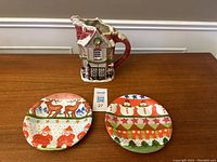 Two Christmas themed plates with colorful holiday scenes and a ceramic house-shaped jug on a wooden table.