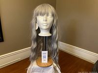 Front view of grey synthetic body wave machine woven wig on a mannequin head showing bangs and wavy texture, displayed indoors on wooden stand.