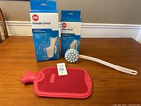 Female and Male Urinals in original boxes, soap/lotion applicator with white handle, and pink hot water bottle on wooden surface.