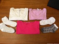 Three knit/jersey scalloped tops folded and two pairs of slipper socks laid out on wooden surface