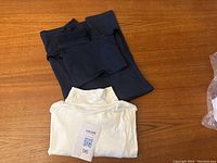 Photo showing folded navy blue top stacked behind white mock turtleneck long sleeve top with a tag showing size XL.