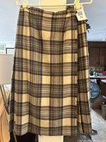 Front view of knee length tartan wool skirt hanging on a hanger showing the plaid pattern in beige and brown.