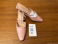 Pair of women's pinky peach slide-on mules placed on wooden surface, showing top and side angles, highlighting croc pattern and kitten heel.