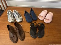 Five pairs of women's shoes on wooden surface: white sneakers, navy slip-ons, pink slides, black sandals, and brown leather boots.