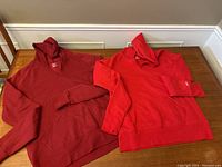 Two women's hooded sweatshirts, one bright red and the other burgundy, laid flat on a wooden surface.