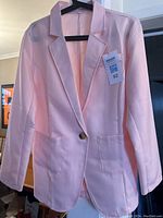 Front view of light pink women's blazer hung on black hanger showing single button closure, patch pockets, and unlined interior.