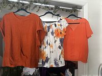Three women's tops hanging on black hangers including orange knit top, white floral top, and orange blouse.