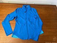 Full view of the blue women's workout jacket with collar and long sleeves laying flat on a wooden table, showing the front zipper and side pockets.