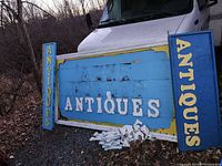 Three blue and yellow wooden signs spelling 'ANTIQUES' including extra wooden letters on the ground in front of the signs outdoors on gravel