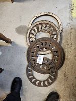 Four cast iron grates stacked on a cement floor showing the different sizes and ornate swirl cut-out designs.