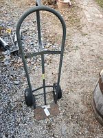 Metal moving hand cart with tubular frame and two wheels shown upright outdoors on gravel and dirt ground. The frame is tubular metal, painted gray with some rust on the platform base. The wheels are black rubber, and one tube is missing.