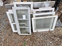 Pile of old wooden window frames stacked outdoors on gravel, featuring mainly white paint and various glass styles.