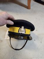 Front view of 1965 RCMP dress cap hat showing black wool felt top, wide yellow band, and silver RCMP badge with crown and horse emblem, and black visor with chin strap.