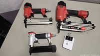 Three air nail tools on a table, two red air nail guns and one silver finish nailer.