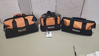 Three Ridgid tool bags of varying sizes shown side by side on a gray floor against a white wall