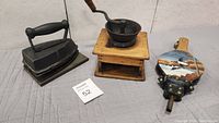 Photo showing antique iron, wooden coffee grinder, and hand-painted bellows arranged on a cloth surface.