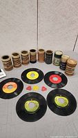 Eight cylindrical Edison wax phonograph rolls of varying labels and conditions alongside five 45 RPM vinyl records with colored center labels and three plastic adapters.