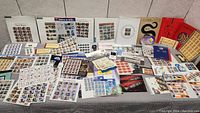 Wide view of the large assortment of full sheets and special edition stamp sheets spread out on a surface. Includes various colorful and theme-based stamp sheets in protective sleeves and loose.