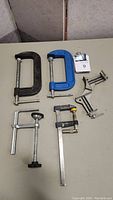 Six clamps of varying sizes and types arranged on a table: two blue and black C-clamps, a silver clamp with black handle, a small yellow and black bar clamp, and two grey specialty clamps with screws for tightening.