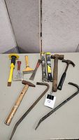 Full lot view of ten construction demo tools on a table including hammers, pry bars, chisels, scrapers, angle grinder, and measuring sticks.