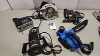 View of all four corded power saws and a plunge router including Black & Decker circular saw, Skil jigsaw, Mastercraft planer, and Black & Decker router, all plugged in with cords.