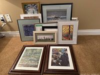 Lot of nine framed art prints, various styles and sizes displayed on brown carpet against a beige wall.