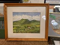 Framed rural landscape painting with green hills, animals, house, and clouds in wooden frame with beige mat.