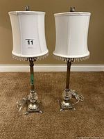 Pair of decorative lamps side by side, white fabric lampshades with decorative tassels, glass and metal bases on carpeted floor.