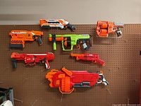 Seven Nerf toys displayed on a brown pegboard, showing the variety and colors of the blasters.
