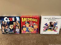 Photo of three board games: Clue, Hedbanz, Trivial Pursuit 2000s boxes side by side on floor.