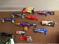 Full view of ten toy blasters displayed on pegboard including seven Nerf blasters and three Super Soakers, various sizes and colors, untested.
