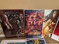 Shows close-up view of Game of Thrones, Avengers: Infinity War, and Wonder Woman posters sealed in plastic with cardboard support.