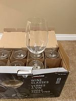 Box containing eight clear Kirkland wine glasses, each separated by cardboard dividers. One wine glass is displayed upright on top of the box showing the entire glass shape.