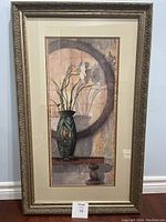 Full view of the framed art showing tall vase with flowers in front of a round mirror and textured background.