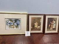 Photo shows three framed art prints on the floor against a wall: two depicting children with dogs and one floral print.