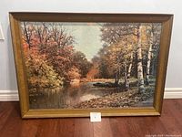 Framed landscape painting showing a calm river with autumn trees in red, orange, yellow, and white foliage. Frame is wooden with a light brown finish showing some wear.
