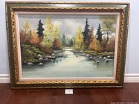 Full frontal photo of the framed signed landscape painting showing autumn river scene with surrounding trees and evergreen background.