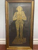 Full view of framed brass rubbing showing a knight in armor on a black background in a gold frame.