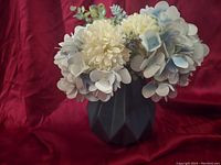 Front view of the artificial floral arrangement showing ivory chrysanthemums, pale blue hydrangeas, green and blue accent foliage in a faceted matte black vase placed against a red fabric background.