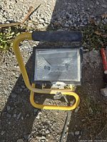 Yellow tubular-framed construction light on ground, with black foam handle and textured glass lens, photographed in natural light.