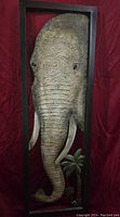 Framed 3D sculpture of an elephant head with tusks and textured surface, including some foliage detail at the bottom in a tall rectangular frame.