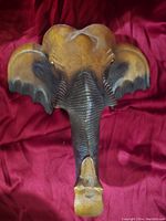 Front view of carved wooden elephant head wall hanging showing detailed trunk and ears on red fabric background.